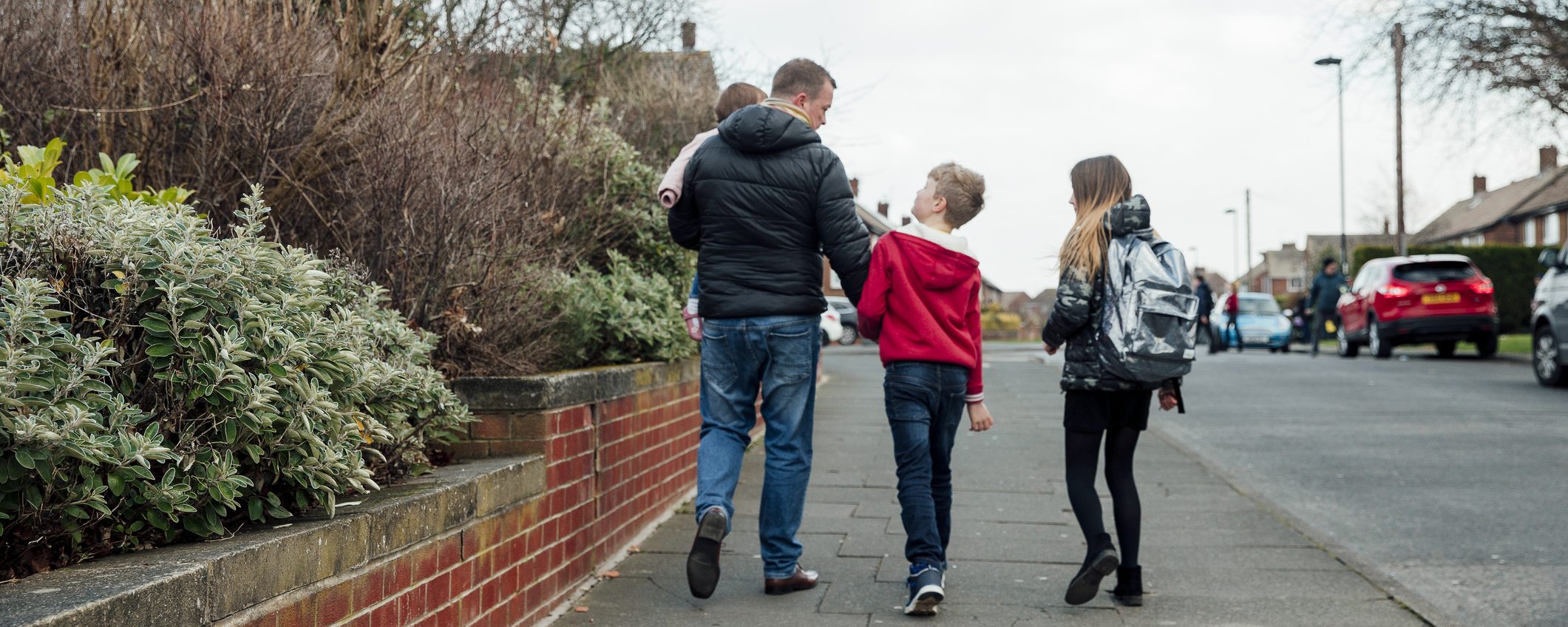 A father holding his toddler and walking with two young children down a neighbourhood path, walking home from school.