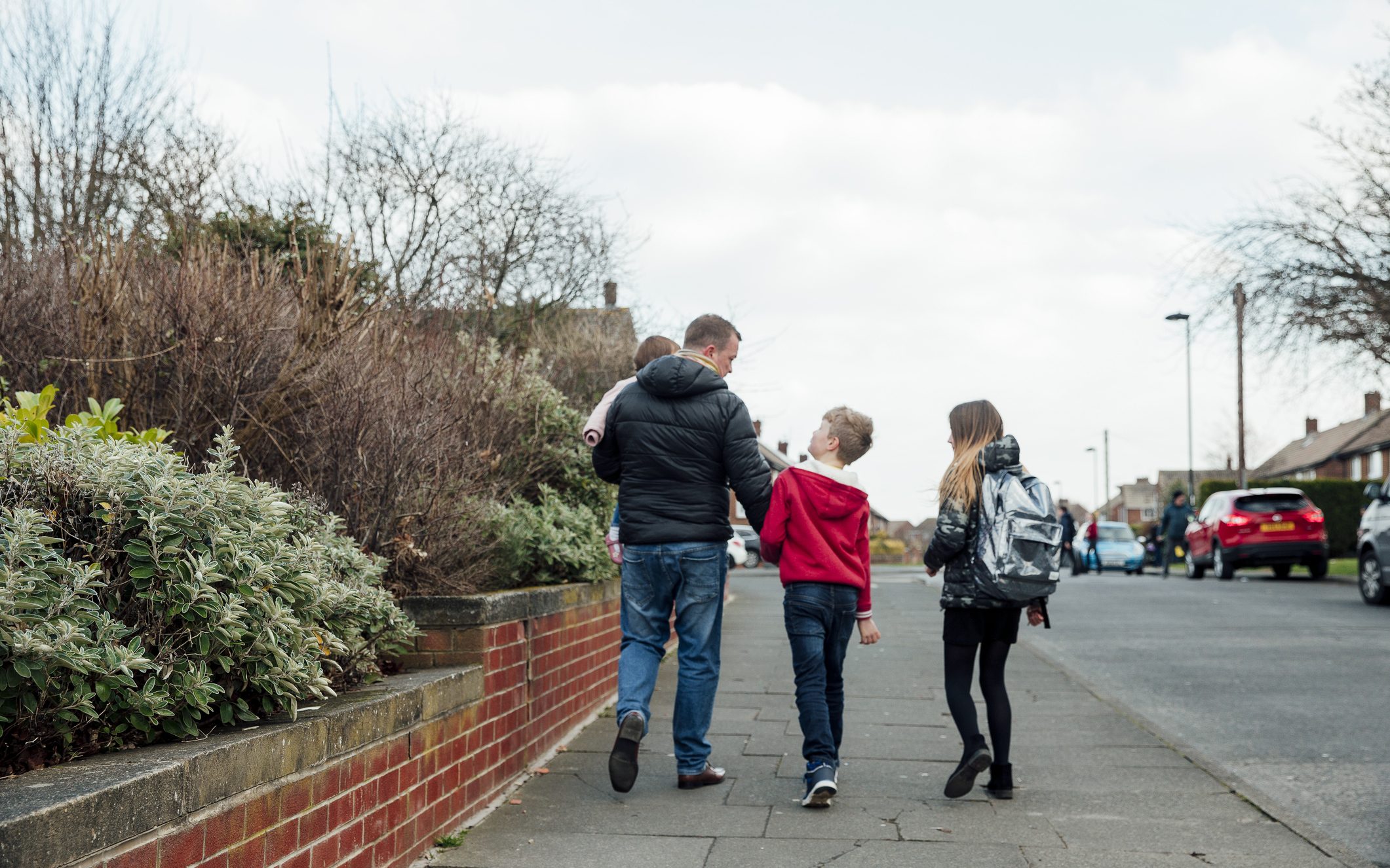 A father holding his toddler and walking with two young children down a neighbourhood path, walking home from school.