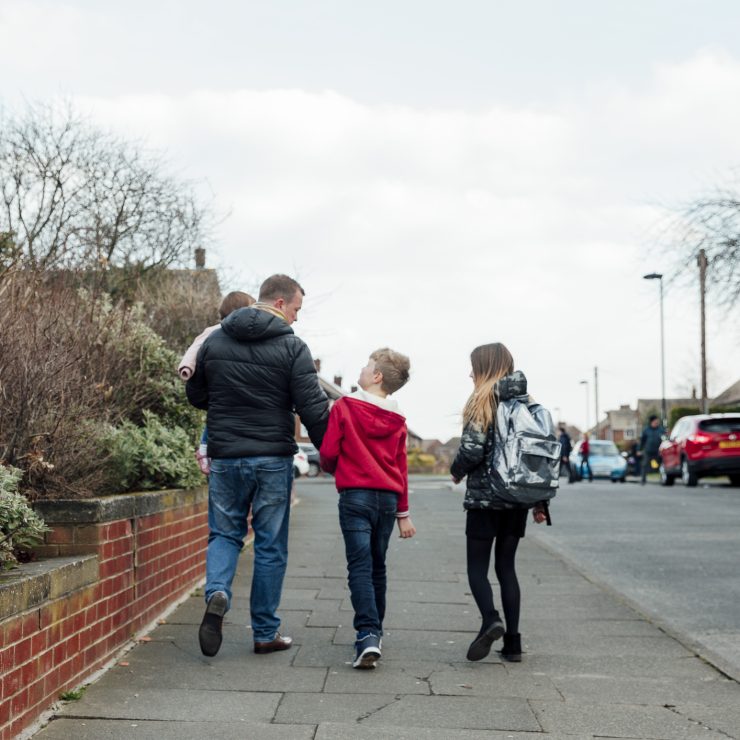 Rear view of a mature man walking down the street with his children. He is holding his sons hand and carrying his baby daughter. A father holding his toddler and walking with two young children down a neighbourhood path, walking home from school.