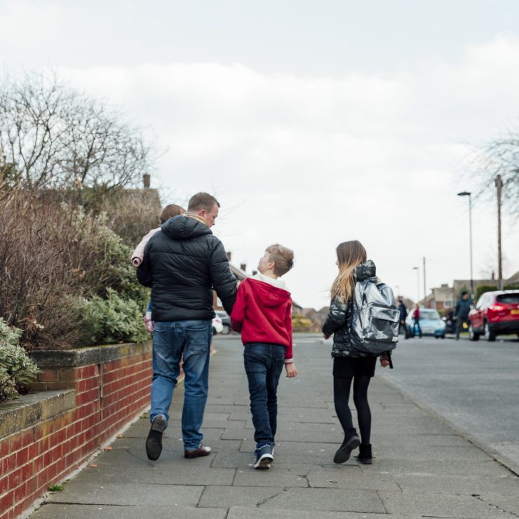 Rear view of a mature man walking down the street with his children. He is holding his sons hand and carrying his baby daughter. A father holding his toddler and walking with two young children down a neighbourhood path, walking home from school.