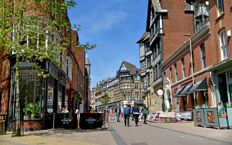 A paved path in Leicester city centre that people re walking down, with shops and public information signage.