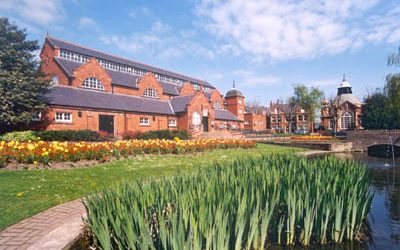Red brick museum building beside a pond and colourful flower beds in a landscaped garden setting