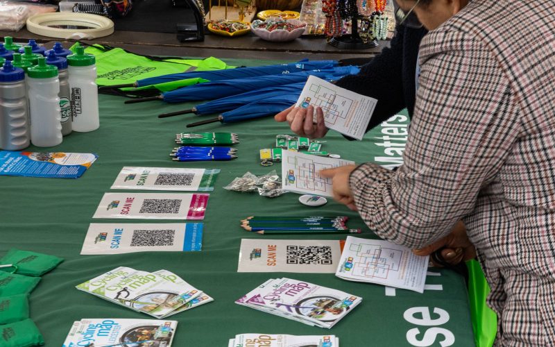 People discussing routes on a Choose How You Move stall, with maps, pencils, bags, umbrellas, and leaflets spread out on the table. For communities and business engagement.