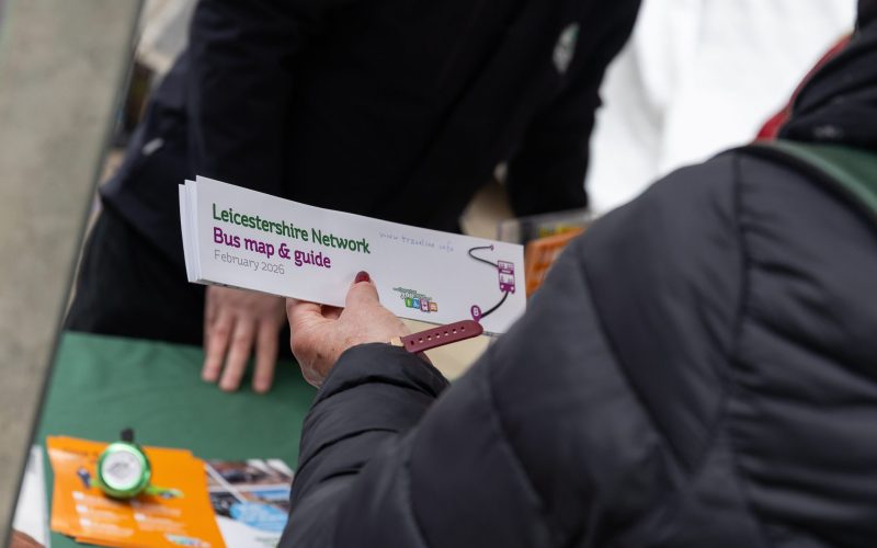 A person holding a bus guide leaflet in her hand, in front of a green-clothed stall. For communities, Leicestershire businesses, public transport.