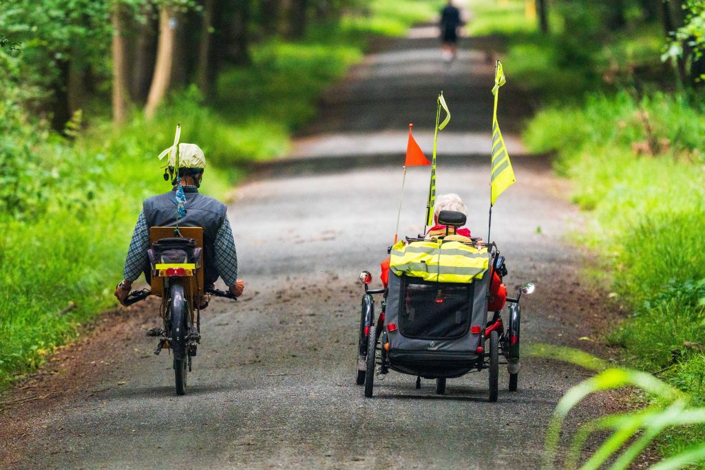 two people cycle down a country path in adapted cycles. The person on the right has red and yellow flags attached to their transport. Showing adaptive bikes and scooters for accessible transport options.