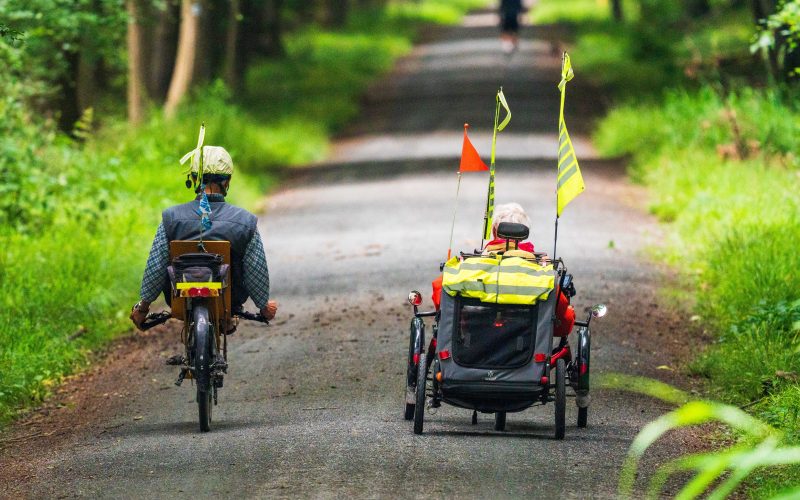 two people cycle down a country path in adapted cycles. The person on the right has red and yellow flags attached to their transport. Showing adaptive bikes and scooters for accessible transport options.