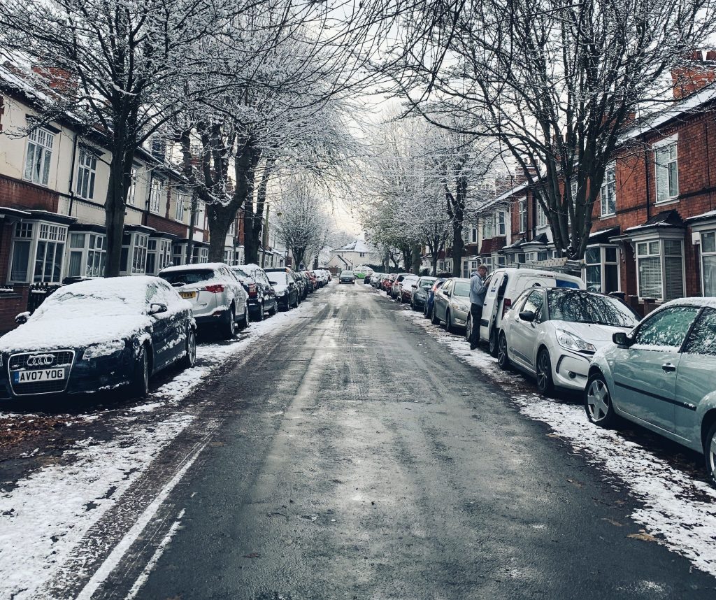 Snow-covered street in Leicester with parked cars and leafless trees during winter. For driving, car sharing, and road safety.