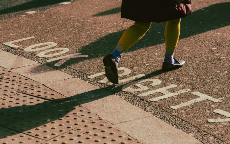 A person in yellow tights with a red skirt crossing at a pedestrian crossing. The road has the words "Look right" and an arrow pointing right painted on in white. For driving, walking, road safety and schools.