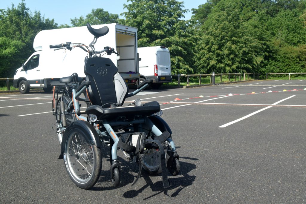 an adapted trike with a forward-facing seat and foot rests, parked in a car park, in front of a white van and white lorry. For adaptive bikes and scooters and adaptive cycling.