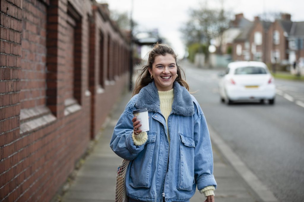 A medium close-up of a young woman wearing a denim jacket walking towards Work. She is carrying a disposable coffee cups and is smiling as she walks towards the town centre she's also carrying a bag over his shoulder as she walks down the road. A woman in a blue jacket holding a coffee and walking to work. For commuting, Leicestershire Businesses, business programme.