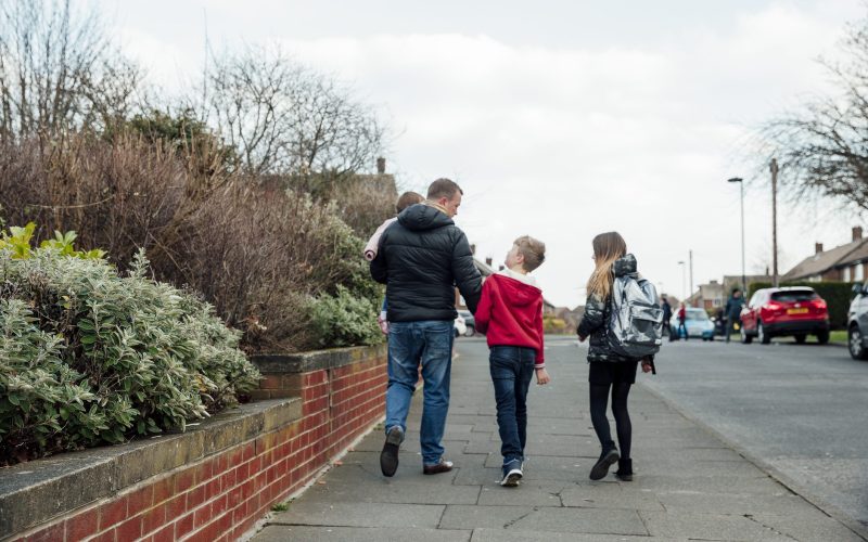 A father holding his toddler and walking with two young children down a neighbourhood path, walking home from school.