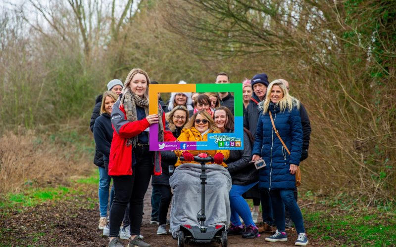 A group people in a wooded area pose with a cardboard frame promoting Choose How You Move