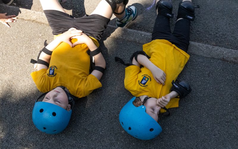 Two children laying down on the school playground wearing helmets and knee pads.