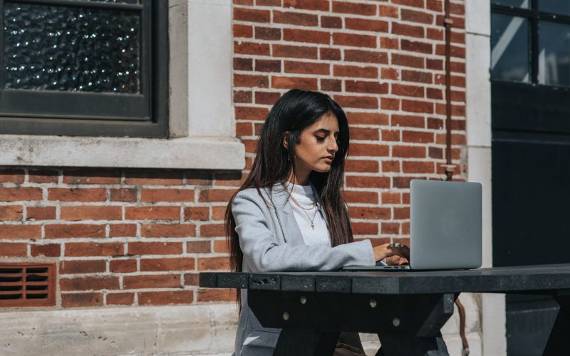 A woman sits at a bench on a sunny day and works from her laptop. A commuter. For businesses, Leicestershire Business programme.
